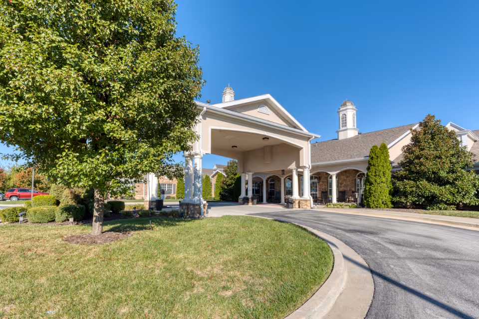 The front exterior of a senior living facility with a covered entrance supported by columns. The building has a beige facade with stone accents and a cupola on the roof. There is a curved driveway leading to the entrance, green grass, and well-maintained bushes and trees under a clear blue sky.