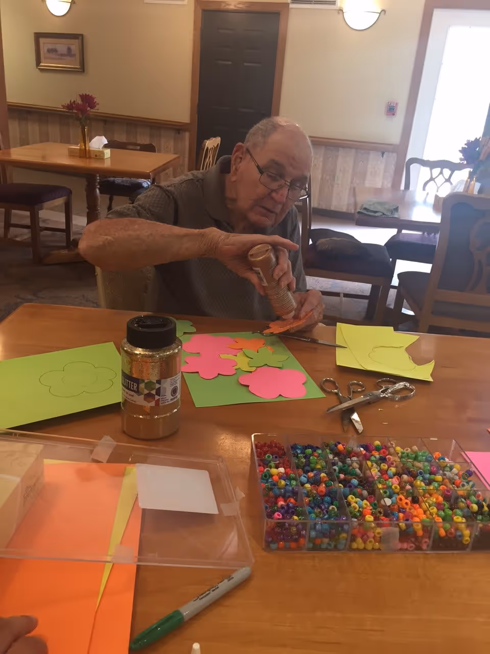 An elderly man sitting at a table in a well-lit room, engaged in a craft activity. He is applying glue to colorful paper cutouts shaped like flowers. The table is covered with various craft supplies including scissors, a container of glitter, colorful beads, and sheets of colored paper. The background shows other tables and chairs in the room with soft lighting and a door.