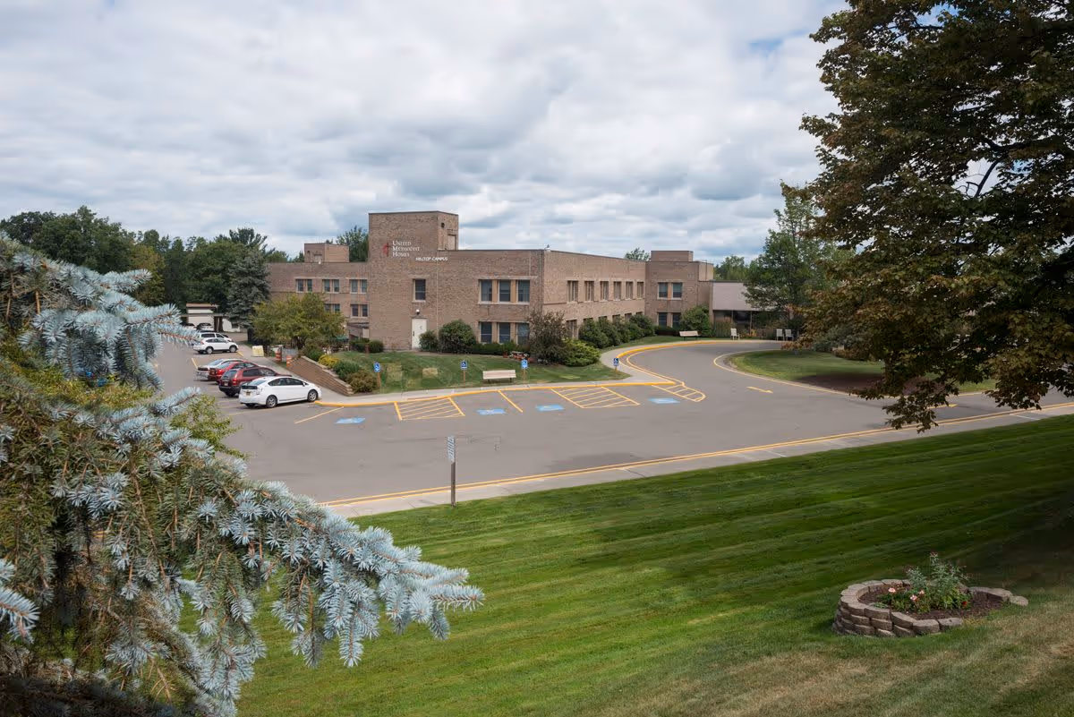 Exterior view of United Methodist Homes Hilltop Campus, a multi-story brick building surrounded by a parking lot with several cars, green lawns, trees, and a partly cloudy sky.