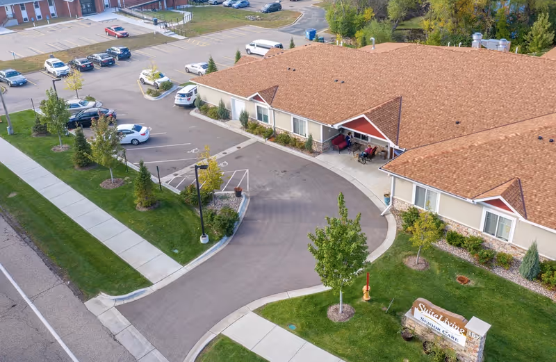 Aerial view of Suite Living Senior Care - Roseville showing a single-story building with a brown shingle roof, surrounded by a parking lot with several cars, green lawns, trees, and sidewalks. There is a sign near the entrance displaying the facility's name.