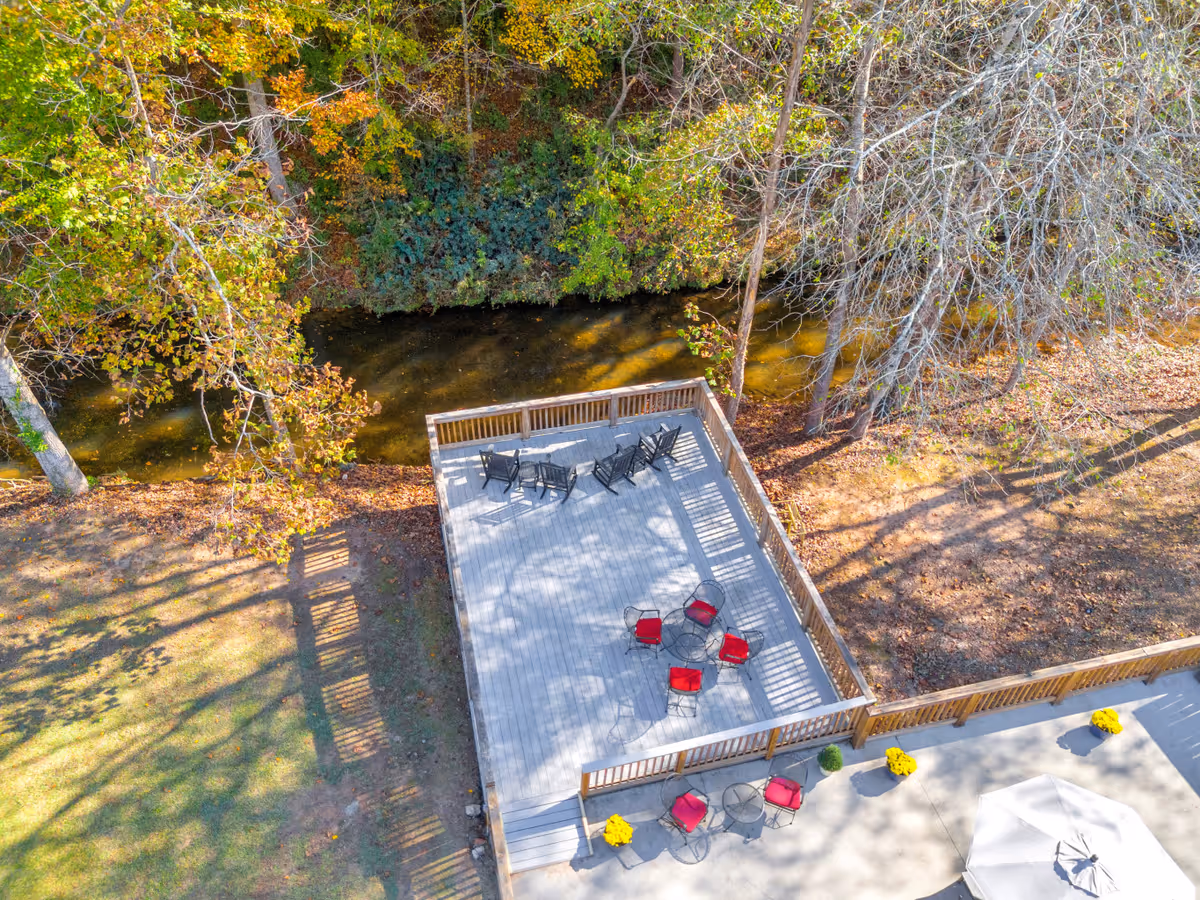 Aerial view of an outdoor wooden deck area with several black and red chairs arranged around small tables. The deck is surrounded by a wooden railing and is adjacent to a small creek with trees showing autumn foliage. There are also yellow potted flowers placed near the deck on a concrete patio with a white umbrella.