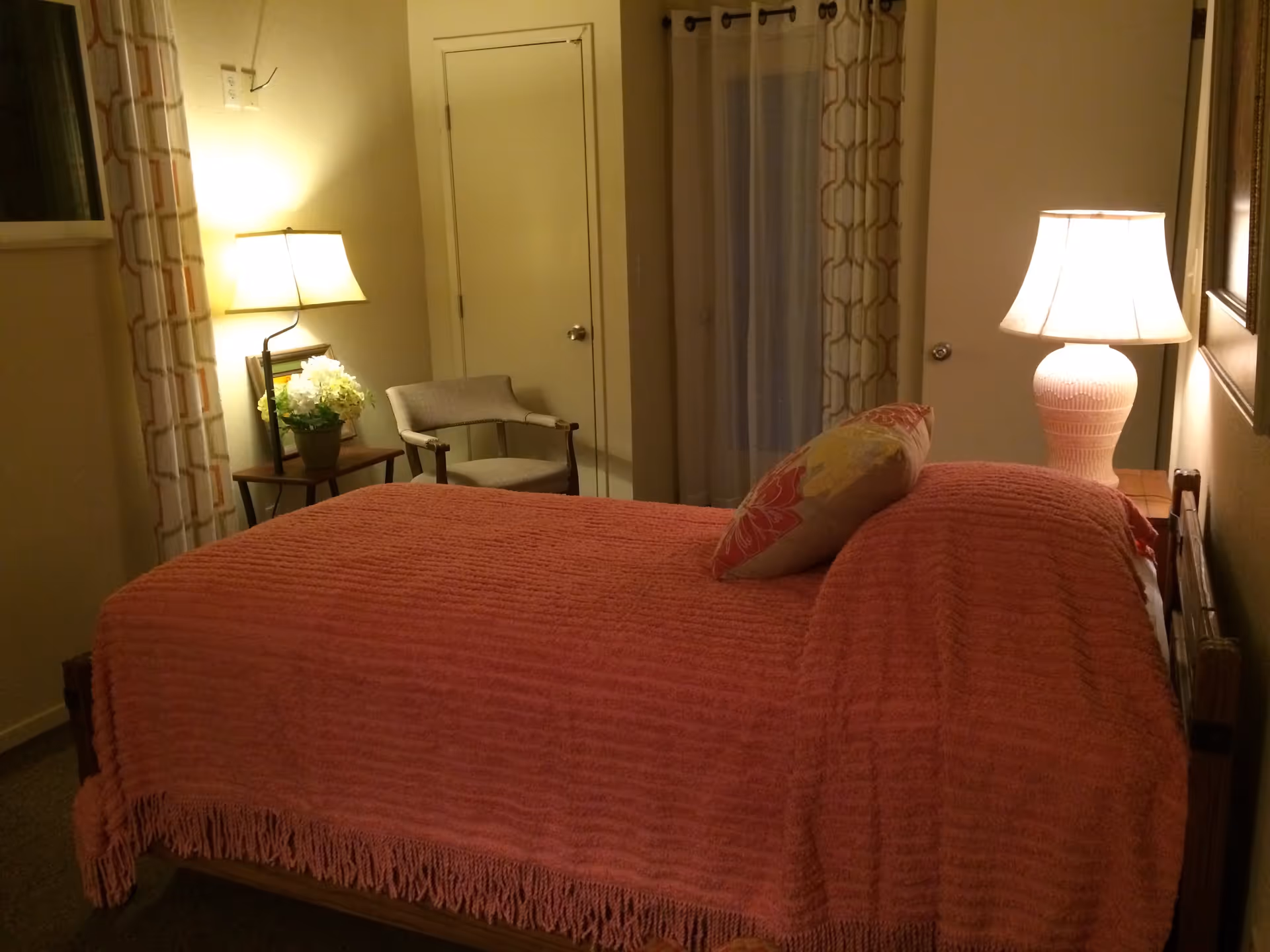 A cozy bedroom with a bed covered in a textured pink blanket and a decorative pillow. There is a wooden nightstand with a white lamp on the right side of the bed. On the left side, there is a small table with a potted plant and a lamp, next to a cushioned chair. The room has light-colored walls, a door, and windows with patterned curtains.