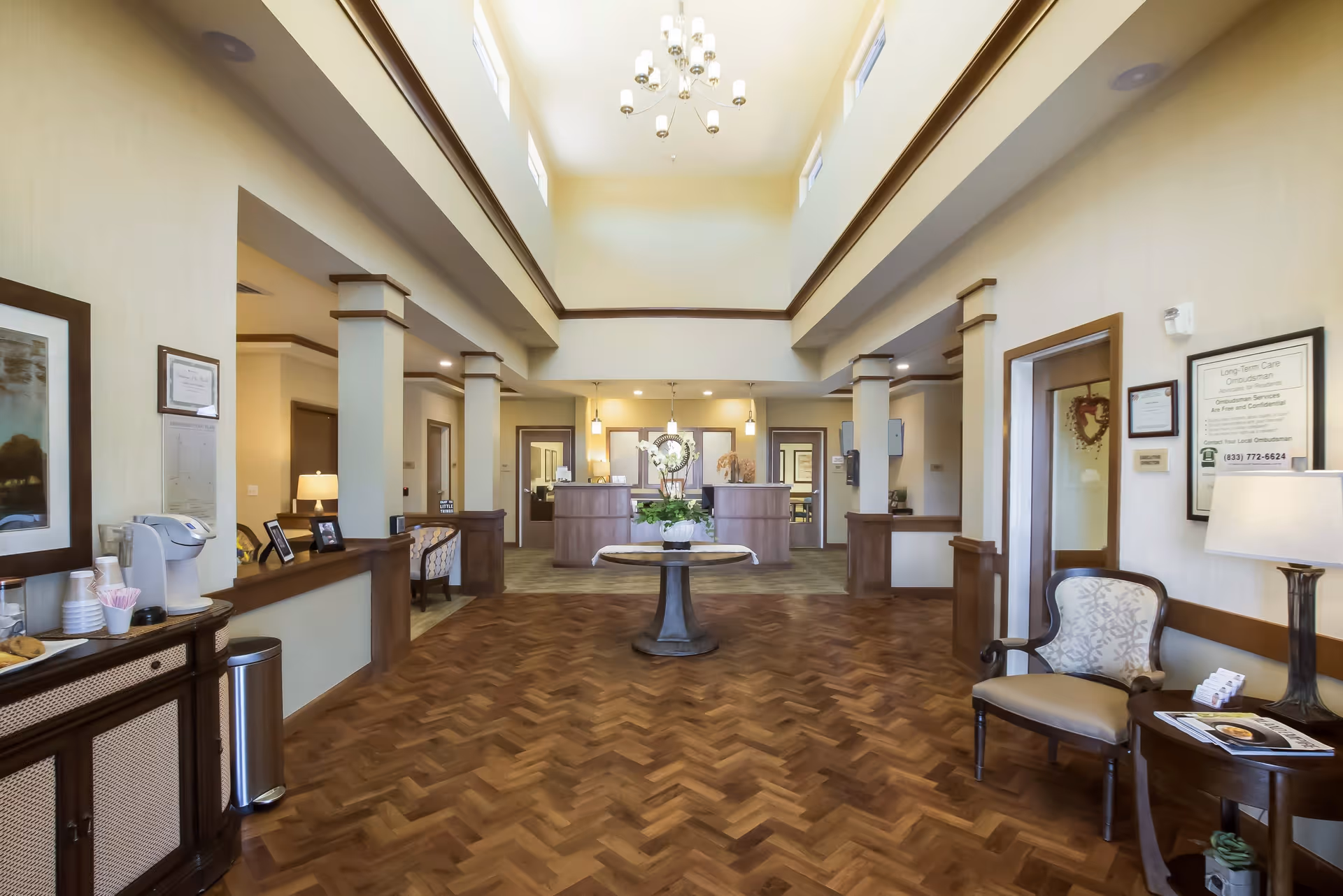 Spacious and well-lit reception area of a senior living facility with a high ceiling, chandelier, wooden flooring, and a round table with a flower arrangement in the center. The reception desk is visible in the background with chairs and framed certificates on the walls.
