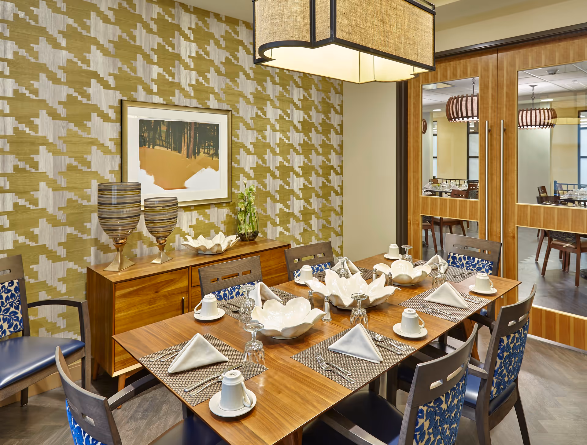 A neatly set dining table in a dining room with patterned wallpaper, wooden cabinets, and decorative accents.
