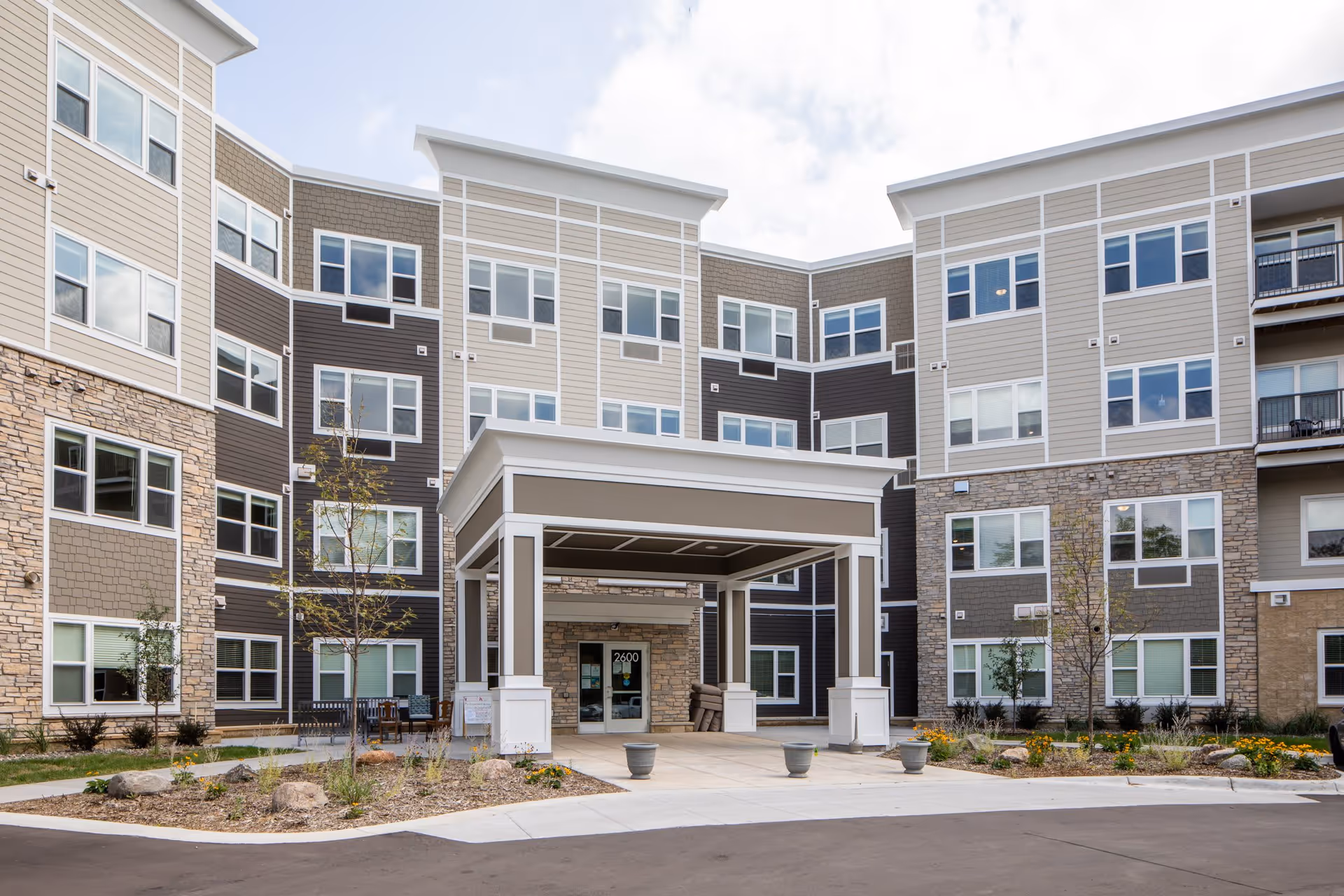 Exterior view of a modern multi-story senior living facility with a covered entrance, landscaped garden beds, and multiple windows.