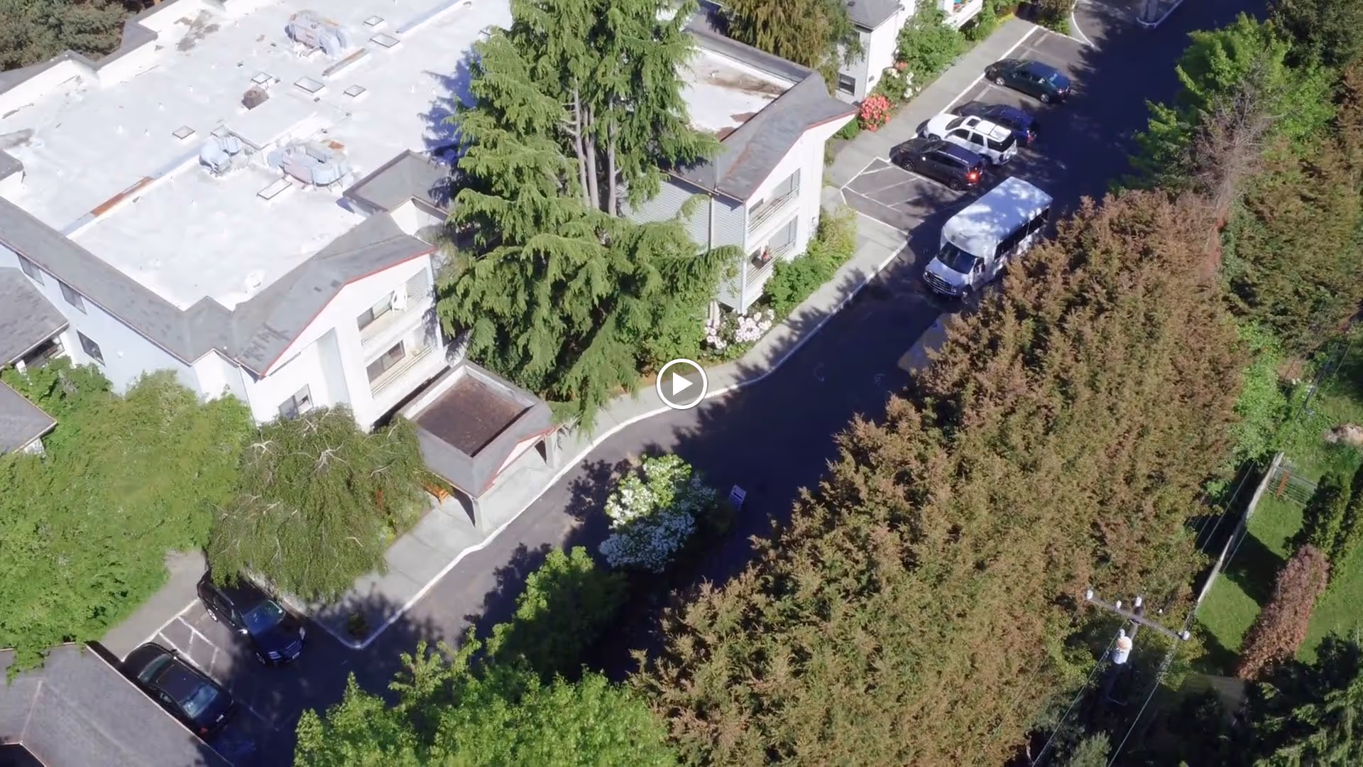 Aerial view of Village Green West Seattle showing a white multi-unit building surrounded by trees and greenery. There is a parking area with several cars and a white van parked along a driveway. The scene is bright and sunny with shadows cast by the trees and building.