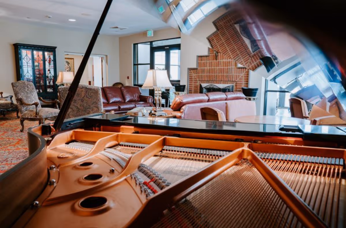 Interior view of a senior living facility lounge area with a grand piano in the foreground, comfortable leather sofas, armchairs, a table lamp, and a brick fireplace in the background.