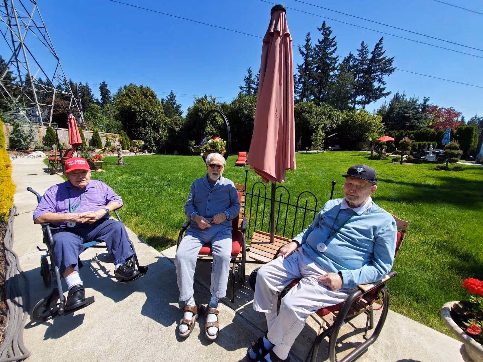 Three elderly men sitting outside on a sunny day in a garden area. Two men are in wheelchairs and one is seated on a chair. There is a closed pink patio umbrella behind them, green grass, trees, and a clear blue sky in the background.