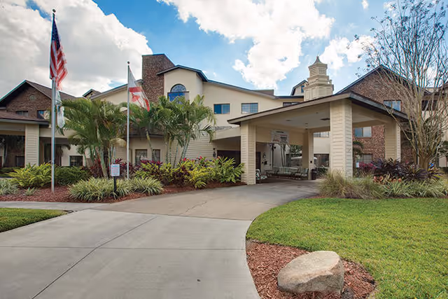 Exterior view of a senior living facility with a covered entrance, landscaped greenery, palm trees, and two flagpoles displaying the American and Florida state flags under a partly cloudy sky.