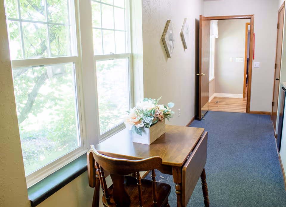 A small wooden table with a floral arrangement in a wooden box sits next to two large windows with a view of green trees outside. A wooden chair is tucked under the table. The room has light-colored walls with two small decorative wall hangings and a blue carpeted floor. An open wooden door leads to another room with wooden flooring.