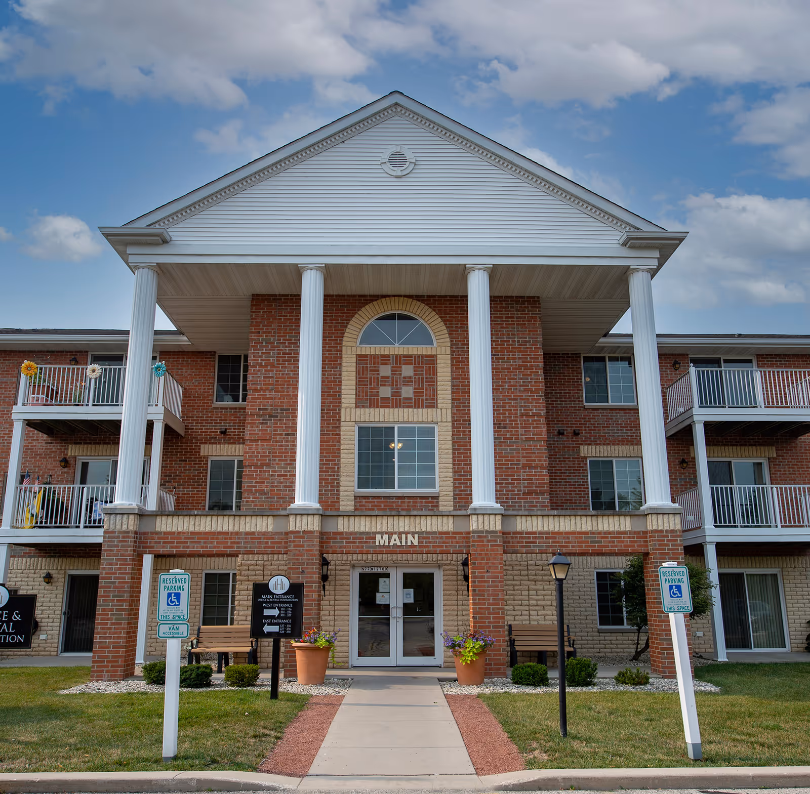Front exterior view of a three-story brick apartment building with white columns and balconies. The entrance has a sign that reads 'MAIN' above double glass doors. There are reserved parking signs for handicapped parking on either side of the walkway leading to the entrance. The sky is partly cloudy.