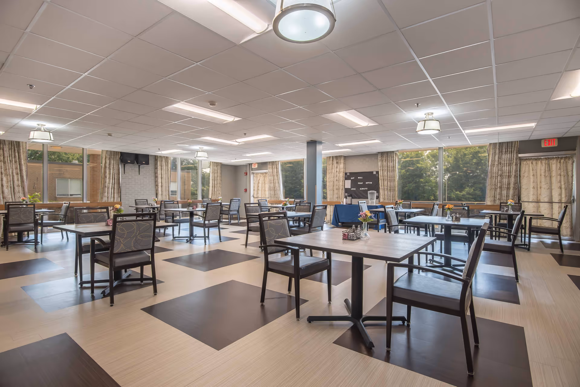 A spacious dining room with multiple tables and chairs arranged neatly. The room has large windows with curtains allowing natural light to enter, and the floor features a checkered pattern with light and dark tiles. There are ceiling lights and a few small flower arrangements on the tables.