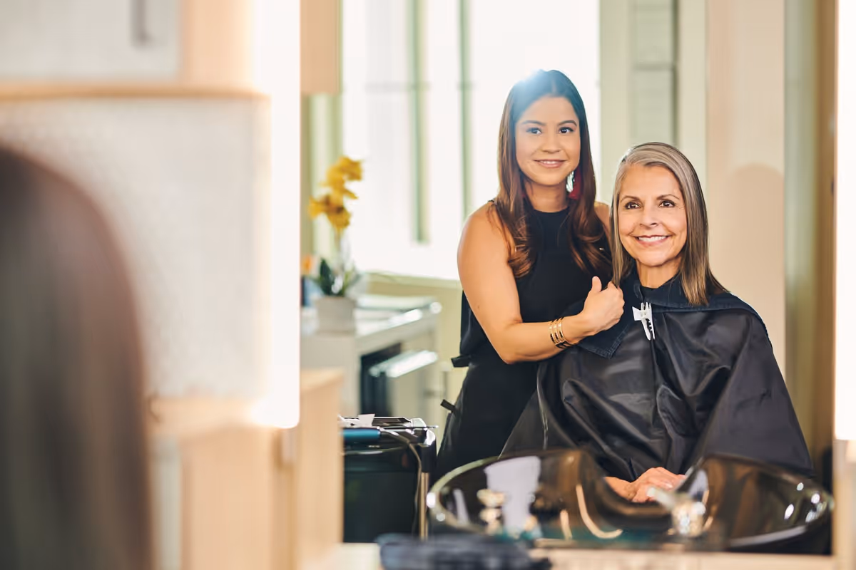 A hairstylist standing behind a seated older woman who is wearing a black salon cape, both smiling and looking into a mirror in a hair salon setting.