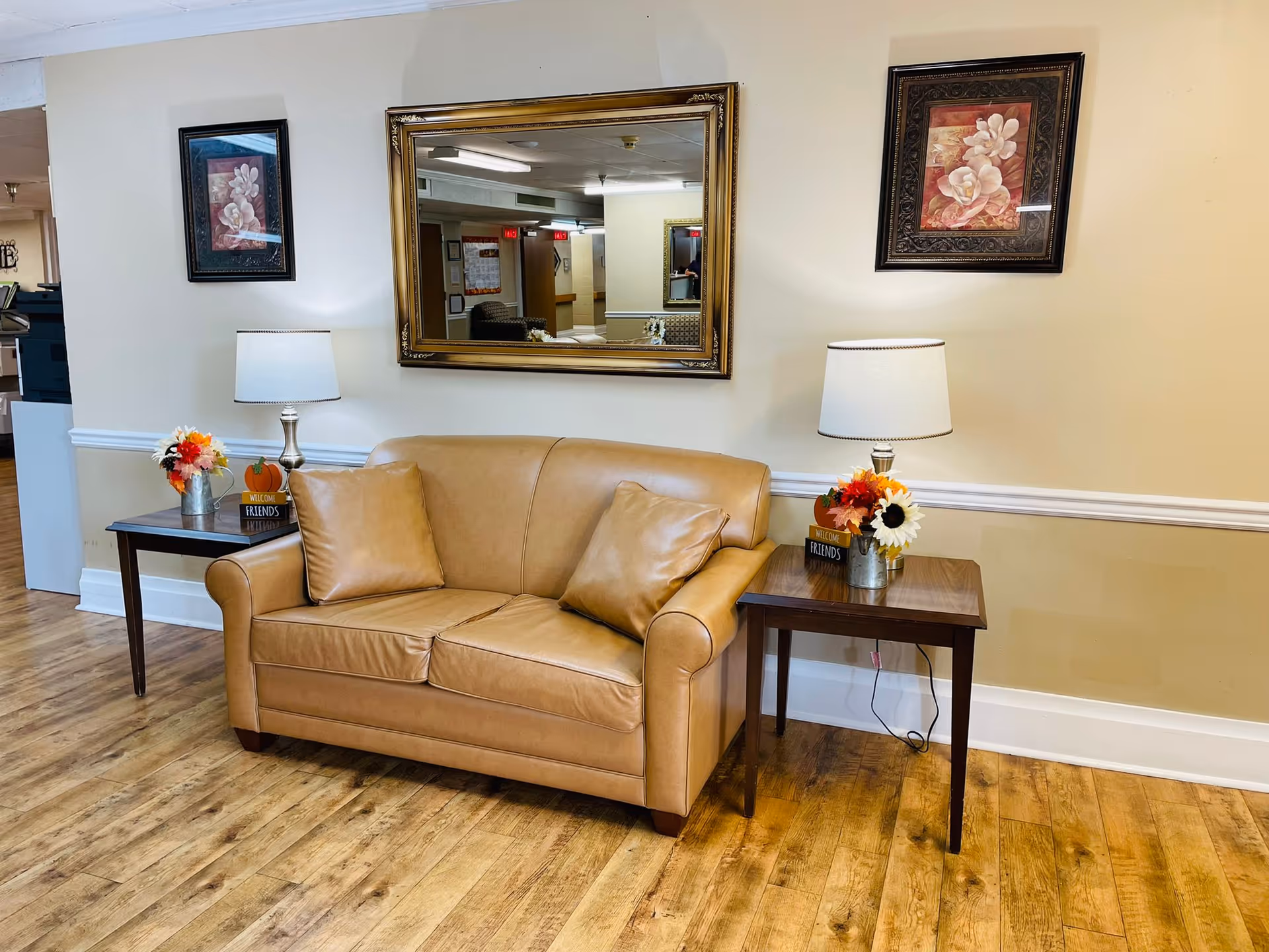 A cozy seating area with a tan leather loveseat flanked by two wooden side tables, each holding a table lamp and a small floral arrangement with a sign that says 'Welcome Friends'. Above the loveseat is a large ornate mirror reflecting part of the room, and on either side of the mirror are framed floral artworks. The floor is wooden, and the walls are painted a light beige color with white trim.