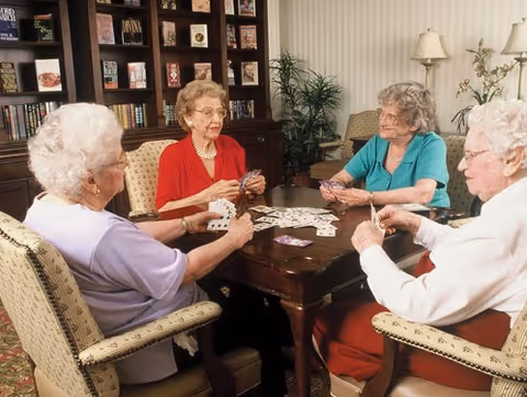 Four elderly women sitting around a wooden table playing cards in a cozy room with bookshelves, plants, and lamps in the background.