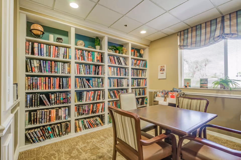 Cozy reading room with wall-to-wall bookshelves, a wooden table surrounded by chairs, and a window letting in daylight.