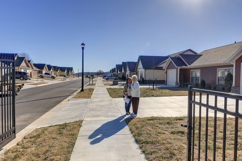 Two elderly women standing on a sidewalk in a residential neighborhood with single-story houses on both sides under a clear blue sky.