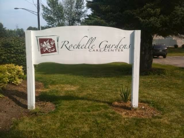 A white wooden sign with a red floral logo and the text 'Rochelle Gardens Care Center' situated on a grassy area with trees and a parked vehicle in the background.