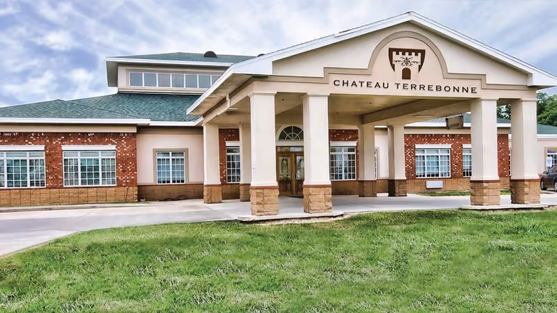 Front exterior view of Chateau Terrebonne Health Care building with a covered entrance supported by columns, brick and beige walls, multiple windows, and a green lawn in front under a partly cloudy sky.