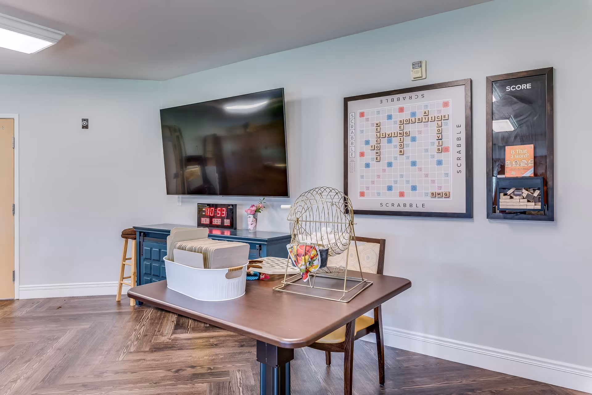 A recreational room with a large flat-screen TV mounted on the wall above a blue cabinet. A digital clock displaying 10:53 is on the cabinet along with a small vase of flowers. On the wall next to the TV is a framed Scrabble board with words related to Solstice Senior Living. Below the board is a table with a bingo cage, a container holding folders, and a chair. The room has wood flooring and light-colored walls.