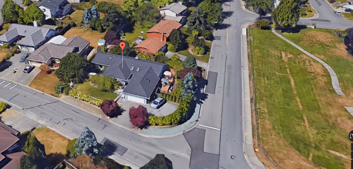 Aerial view of a residential neighborhood showing a single-story house with a dark roof, surrounded by trees and bushes. The house is located at a corner with streets on two sides and a large grassy area nearby.