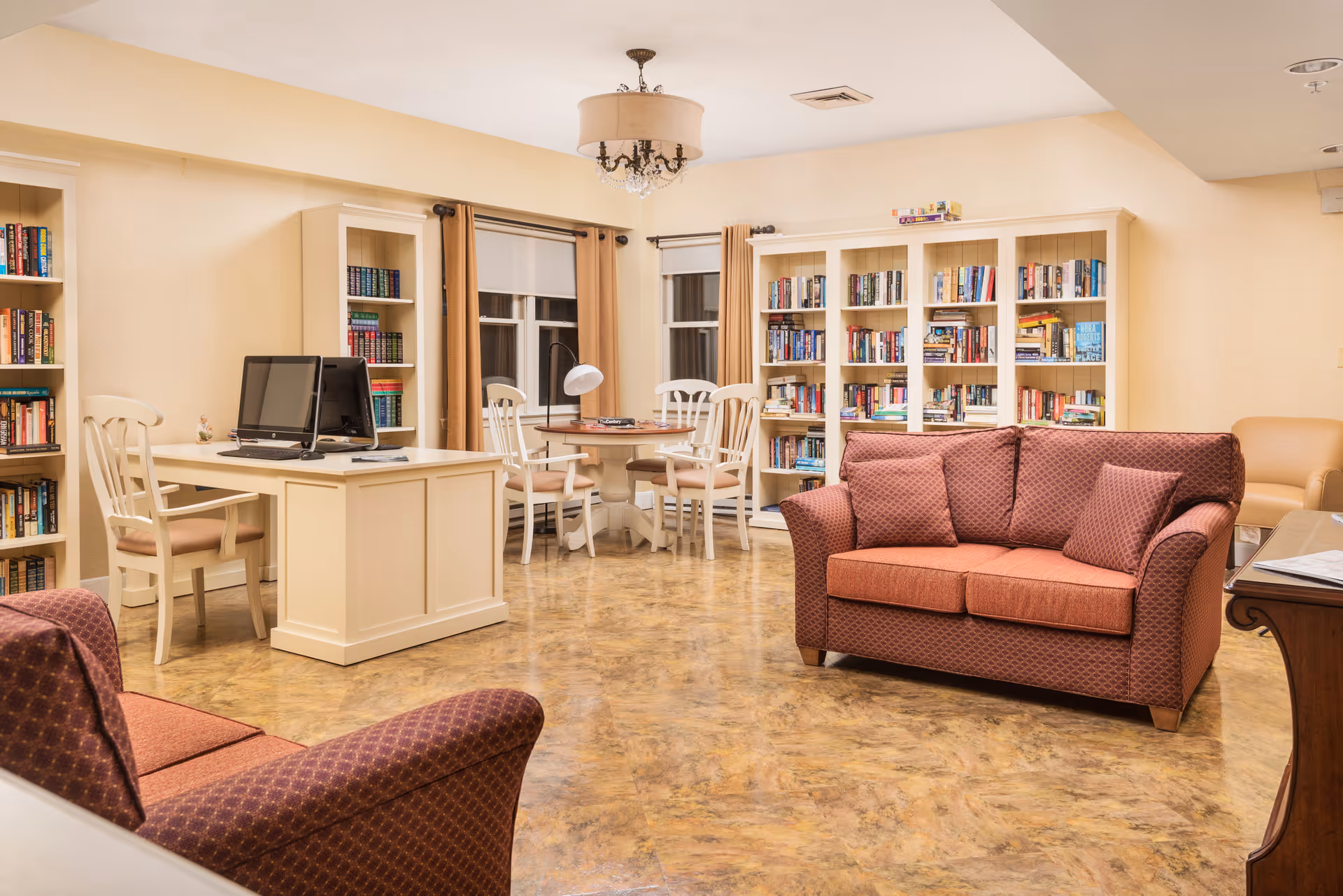 A cozy and well-lit common area featuring a red patterned loveseat and armchair, a white desk with a computer, a round table with four chairs, and multiple bookshelves filled with books. The room has beige walls, large windows with brown curtains, and a chandelier hanging from the ceiling.