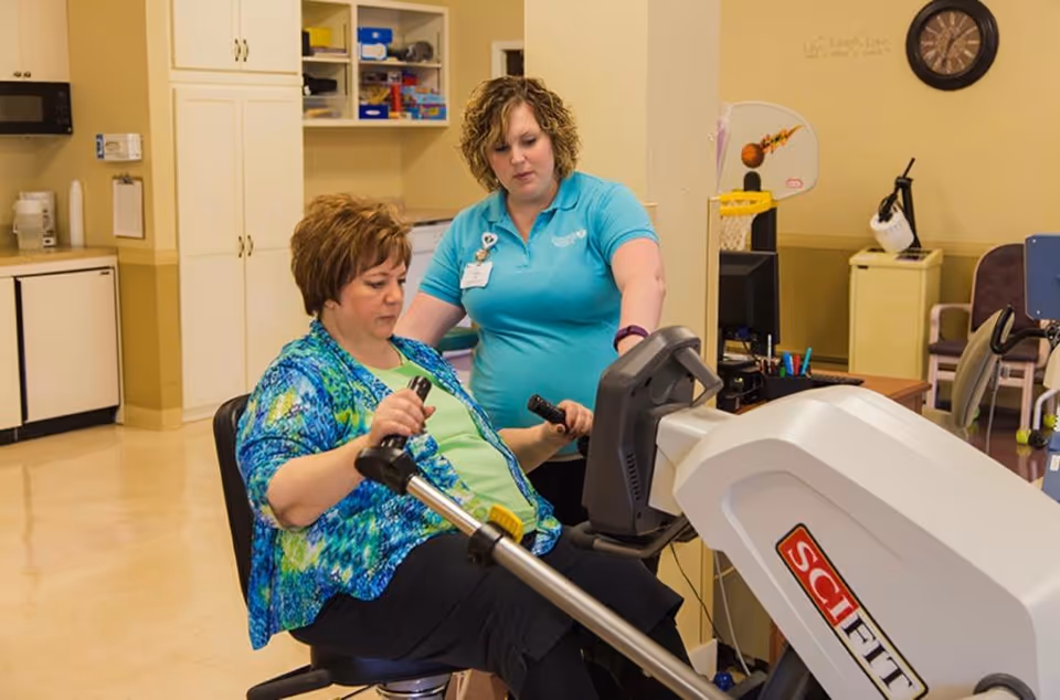 A woman seated on a SciFit exercise machine is being assisted by a staff member wearing a blue polo shirt in a rehabilitation or therapy room with cabinets, a microwave, and chairs in the background.