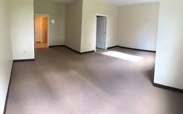 Empty room with beige walls and carpeted floor, two doorways leading to other rooms, and sunlight coming through a window on the right side.