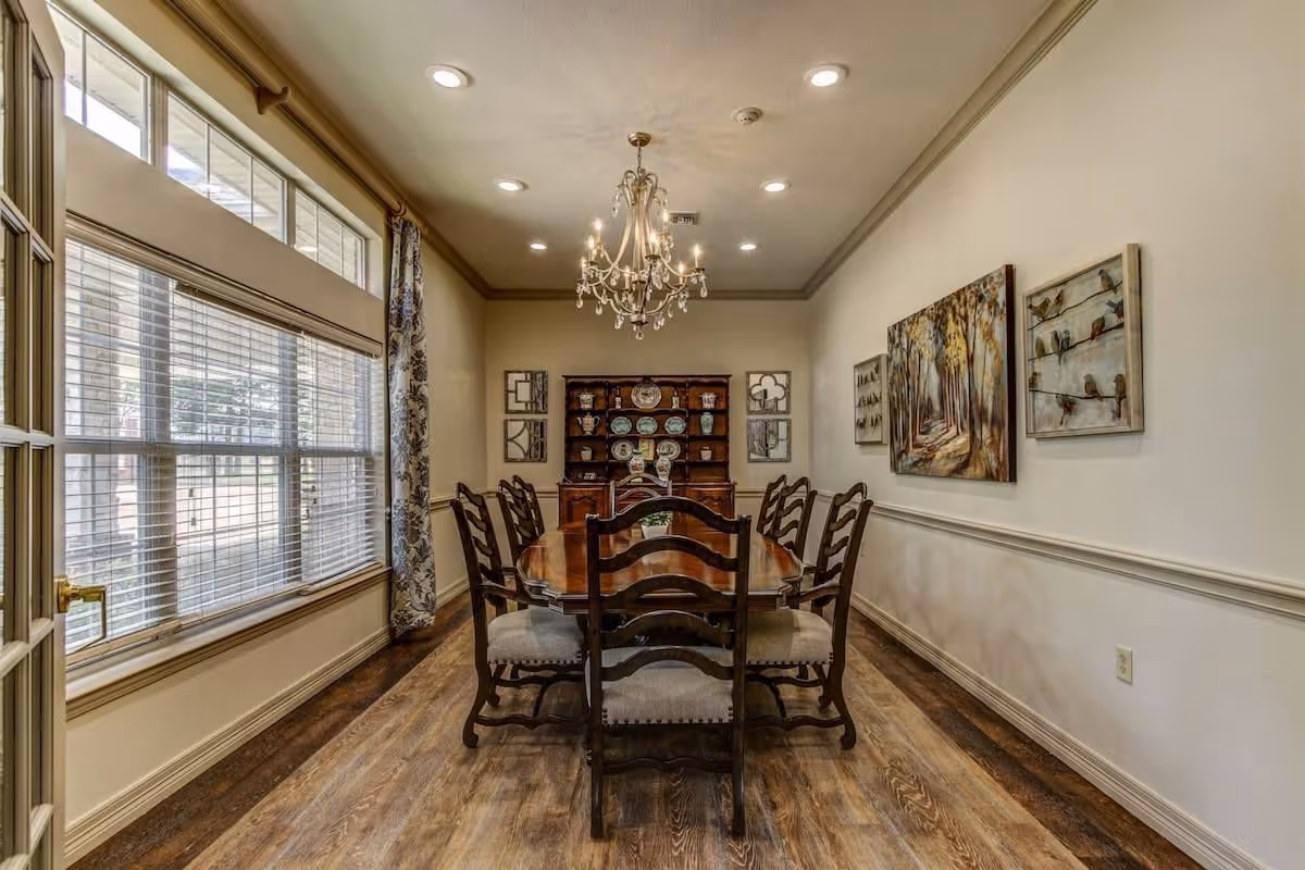Formal dining room with a long wooden table and chairs, a chandelier, a china hutch, and a large window with blinds.