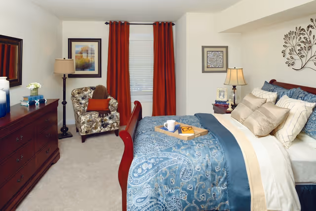 A cozy bedroom with a large bed covered in blue and beige patterned bedding. There is a wooden dresser on the left side with decorative items, a floral armchair with an orange pillow near a window with red curtains, and a nightstand with a lamp and framed photo. The walls are decorated with framed artwork and a metal tree wall sculpture.