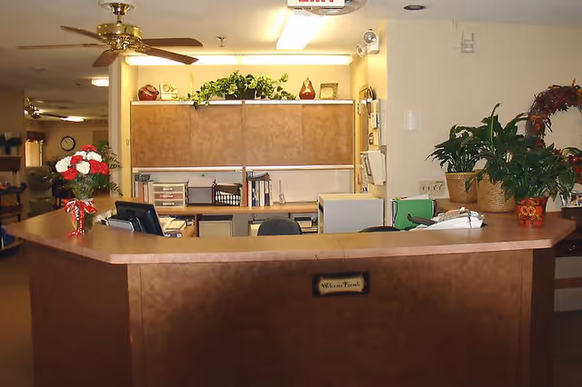Reception desk and office area with computer monitors, potted plants and decorative items in a care facility lobby.