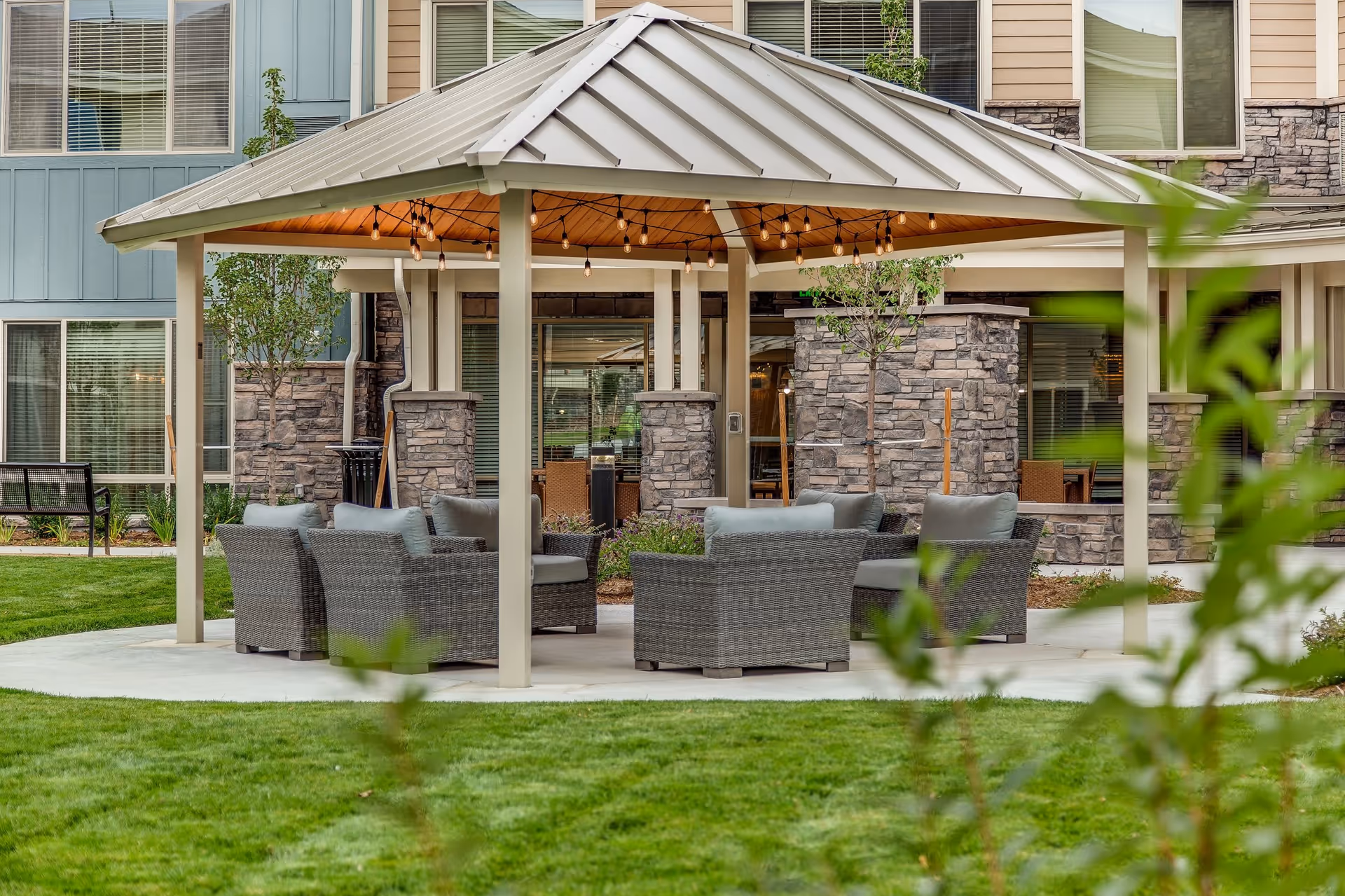 Outdoor seating area under a large gazebo with a metal roof, featuring several cushioned wicker chairs arranged in a circle on a concrete patio, surrounded by green grass and landscaping in front of a building with stone and siding exterior walls and multiple windows.
