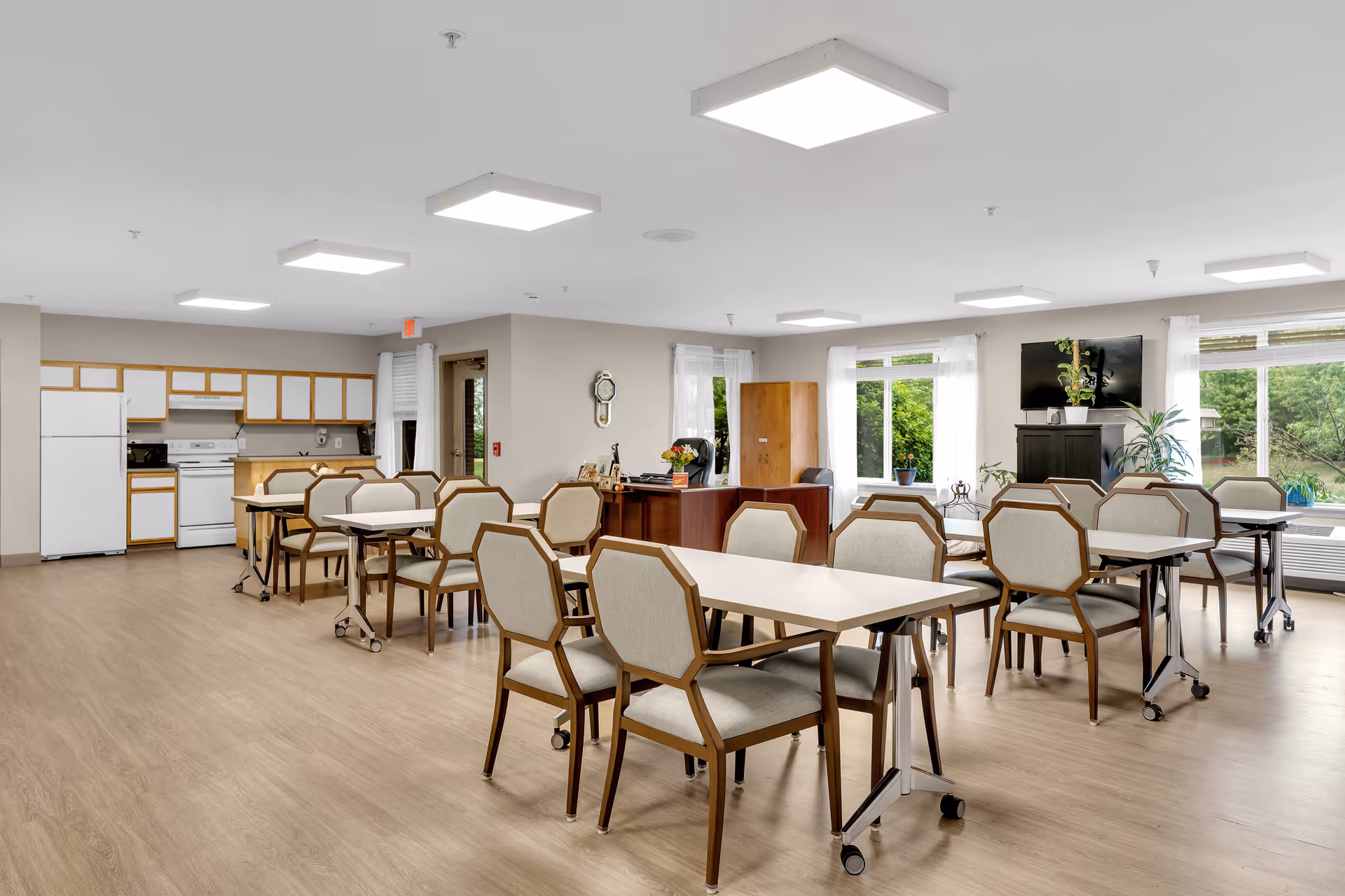 A bright and spacious common area in a senior living facility featuring multiple tables with cushioned chairs on wheels, a kitchenette with white appliances and wooden cabinets, large windows with white curtains letting in natural light, a wooden desk with office supplies, and several potted plants near a mounted flat-screen TV.