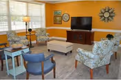 Bright sitting room with patterned armchairs arranged around a central ottoman, a TV on a console, side tables and a large window.
