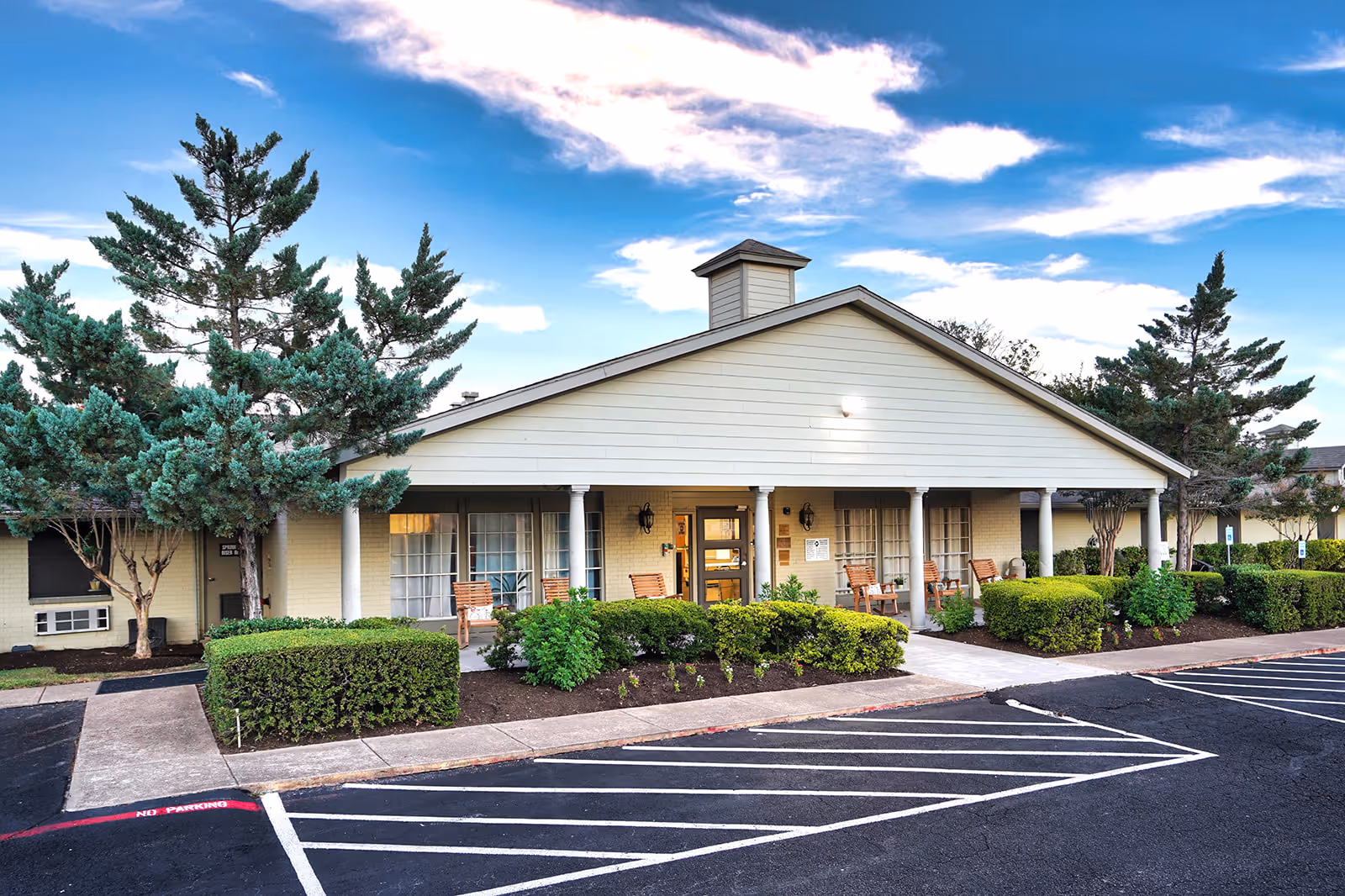 Single-story nursing facility building with a covered front porch, rocking chairs, landscaped shrubs and a parking lot under a blue sky.