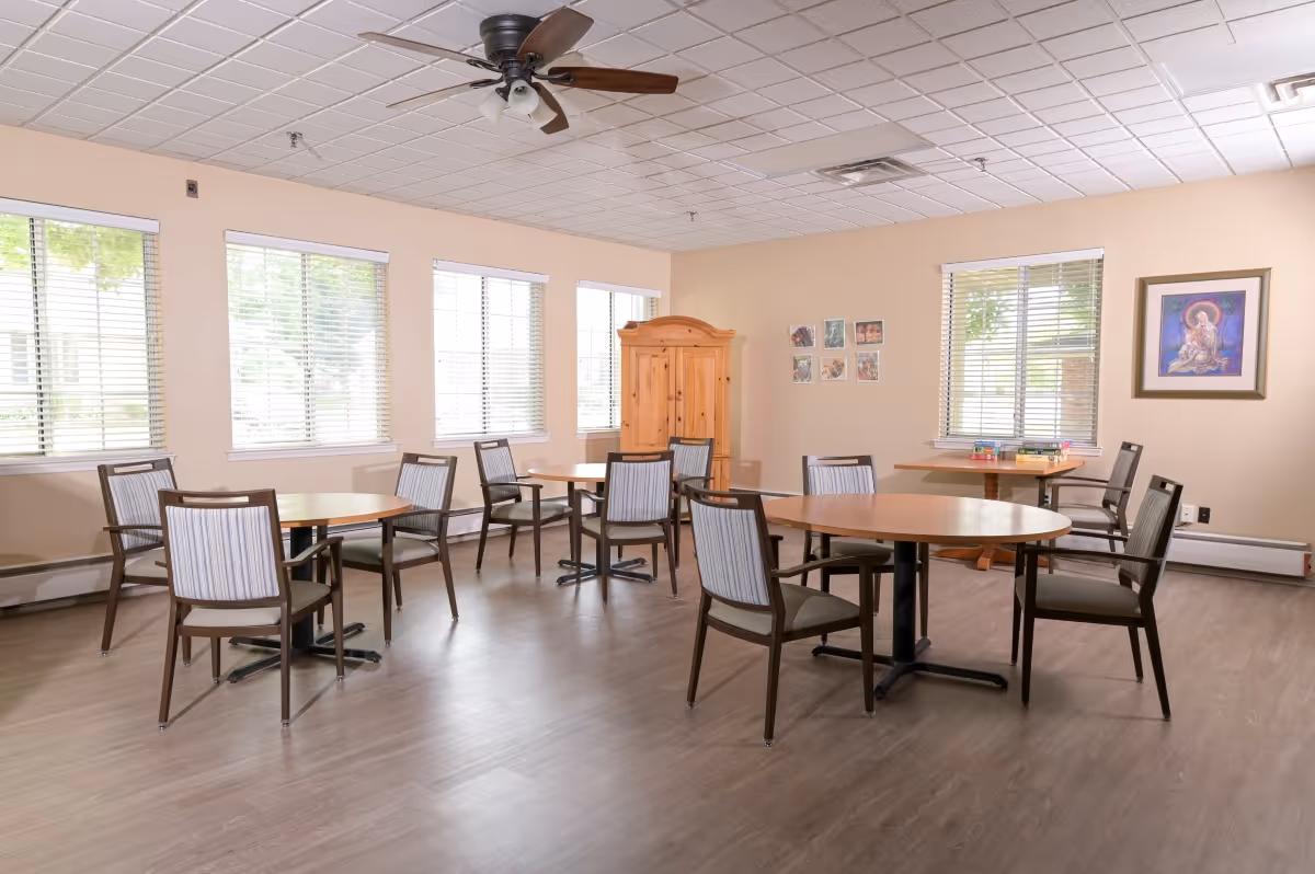 Bright communal dining room with round tables, chairs, large windows, and a ceiling fan.