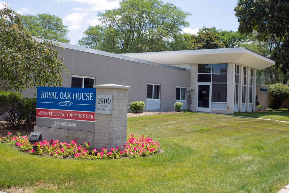 Exterior view of Royal Oak House Assisted Living and Memory Care building with a sign displaying the facility name, services, phone number, and address 1900 North Washington. The building is surrounded by green grass, trees, and flower beds with pink flowers.