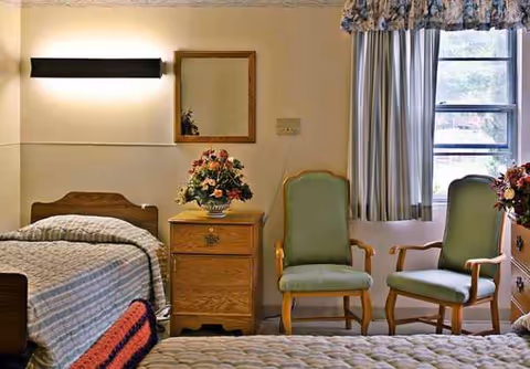 Two beds, a dresser with flowers, two green chairs, and a window in a cozy bedroom.