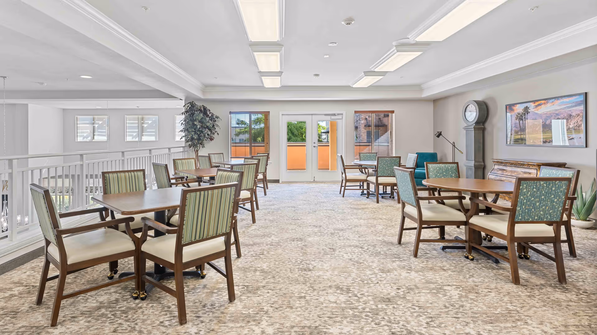 A bright and spacious common area with multiple wooden tables and chairs arranged for seating. The room features large windows and glass doors letting in natural light, a tall grandfather clock, a framed landscape picture on the wall, and a potted plant. The carpet has a neutral pattern, and the ceiling has recessed lighting panels.