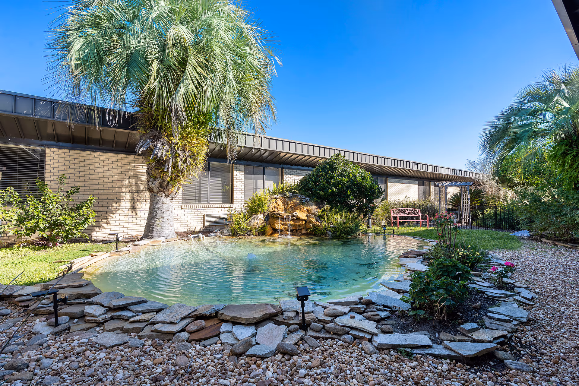 Sunlit courtyard with a small decorative pond, palm trees, and a bench in front of a single-story brick building.