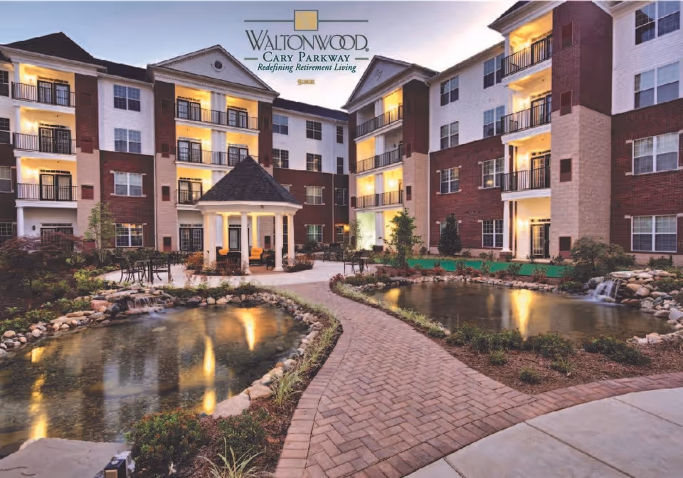 Evening view of Waltonwood Cary Parkway senior living facility showing a courtyard with a brick pathway, two small ponds with waterfalls, a gazebo with seating, outdoor tables and chairs, and a multi-story building with balconies and lit windows in the background.