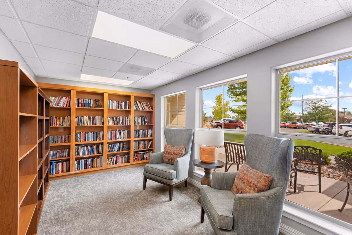 A cozy reading room with two gray upholstered armchairs, each with a patterned cushion, positioned near large windows overlooking an outdoor patio and parking lot. A small round table with a lamp sits between the chairs. Wooden bookshelves filled with books line the wall opposite the windows. The room has a light gray carpet and a white ceiling with recessed lighting.