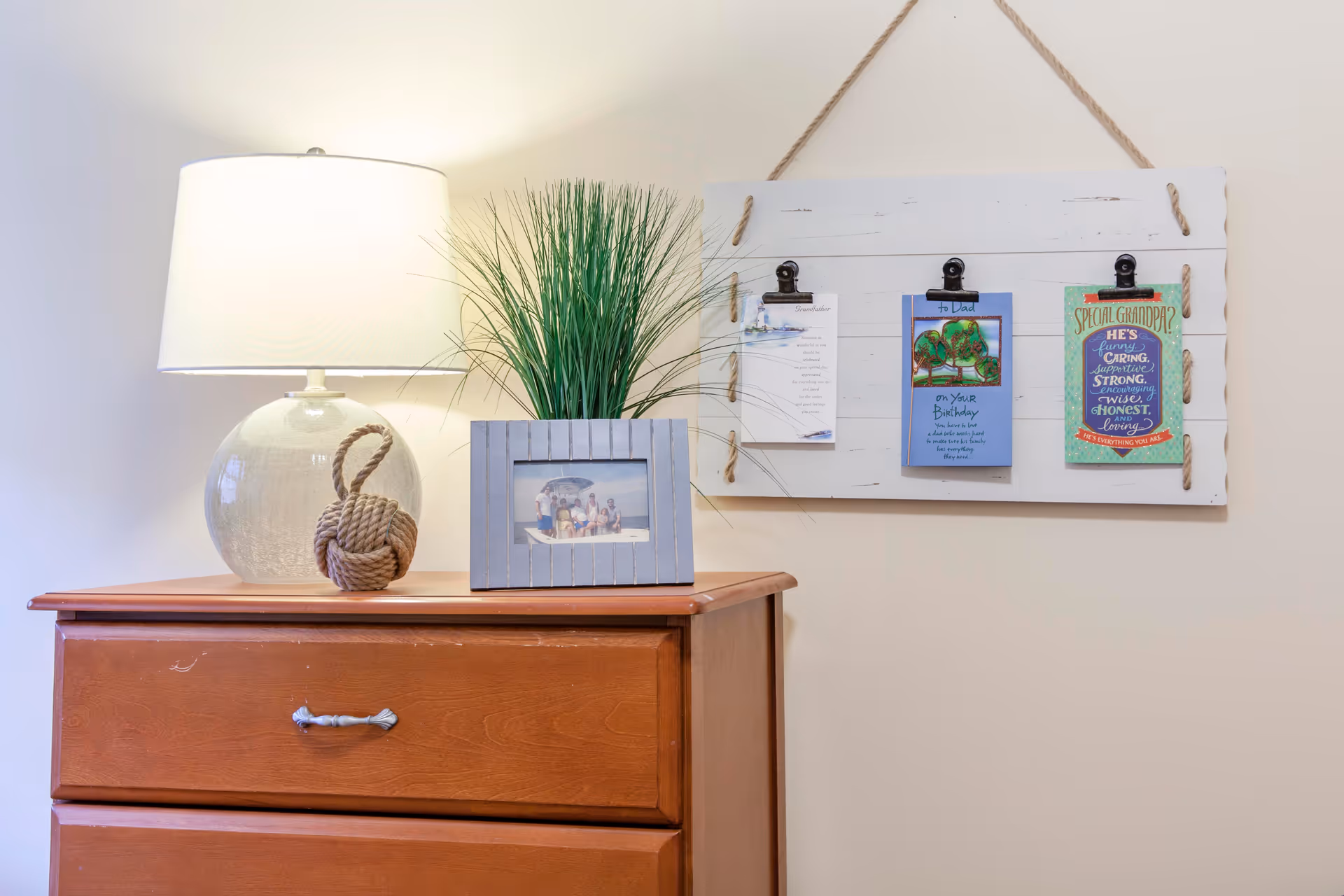 A wooden dresser topped with a lamp, decorative rope knot, framed photo and potted grass, with a white clip-board style wall hanging above.