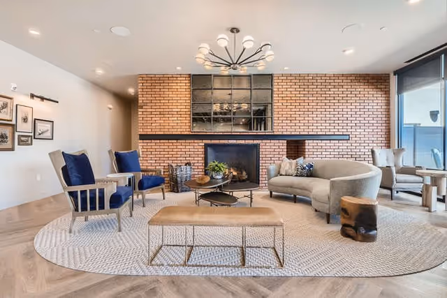 A modern living room with a large circular rug, a curved gray sofa, two blue cushioned armchairs, a wooden bench, and a round coffee table with decorative items. The back wall features a brick fireplace with a large window above it, and there are additional chairs and a small table near a large window with a roller shade.