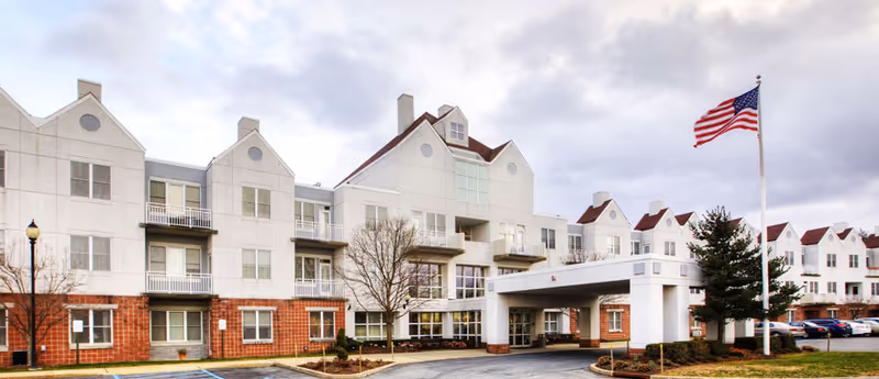 Exterior view of a multi-story senior living facility with white and red brick facade, multiple windows, balconies, an entrance canopy, a flagpole with an American flag, and a parking area in front under a cloudy sky.