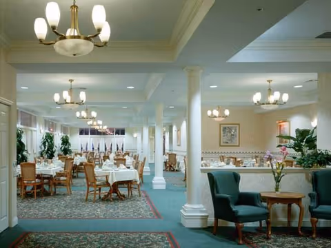A spacious dining room with multiple tables covered in white tablecloths and set with glassware and napkins. Wooden chairs surround the tables. The room features green carpeting with a floral pattern, white columns, and several chandeliers hanging from the ceiling. There are green upholstered armchairs and small wooden tables in the foreground, along with potted plants and framed artwork on the walls.