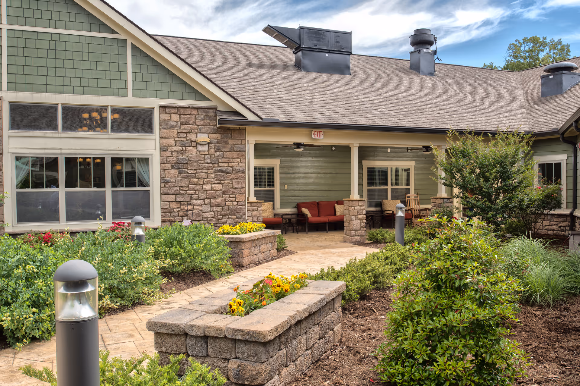 Outdoor patio area at Dominion Senior Living of Crossville featuring a paved walkway, stone planters with flowers, green shrubs, and a covered seating area with red cushioned chairs and a rocking chair. The building exterior has green siding, stone accents, and multiple windows under a sloped roof with vents.
