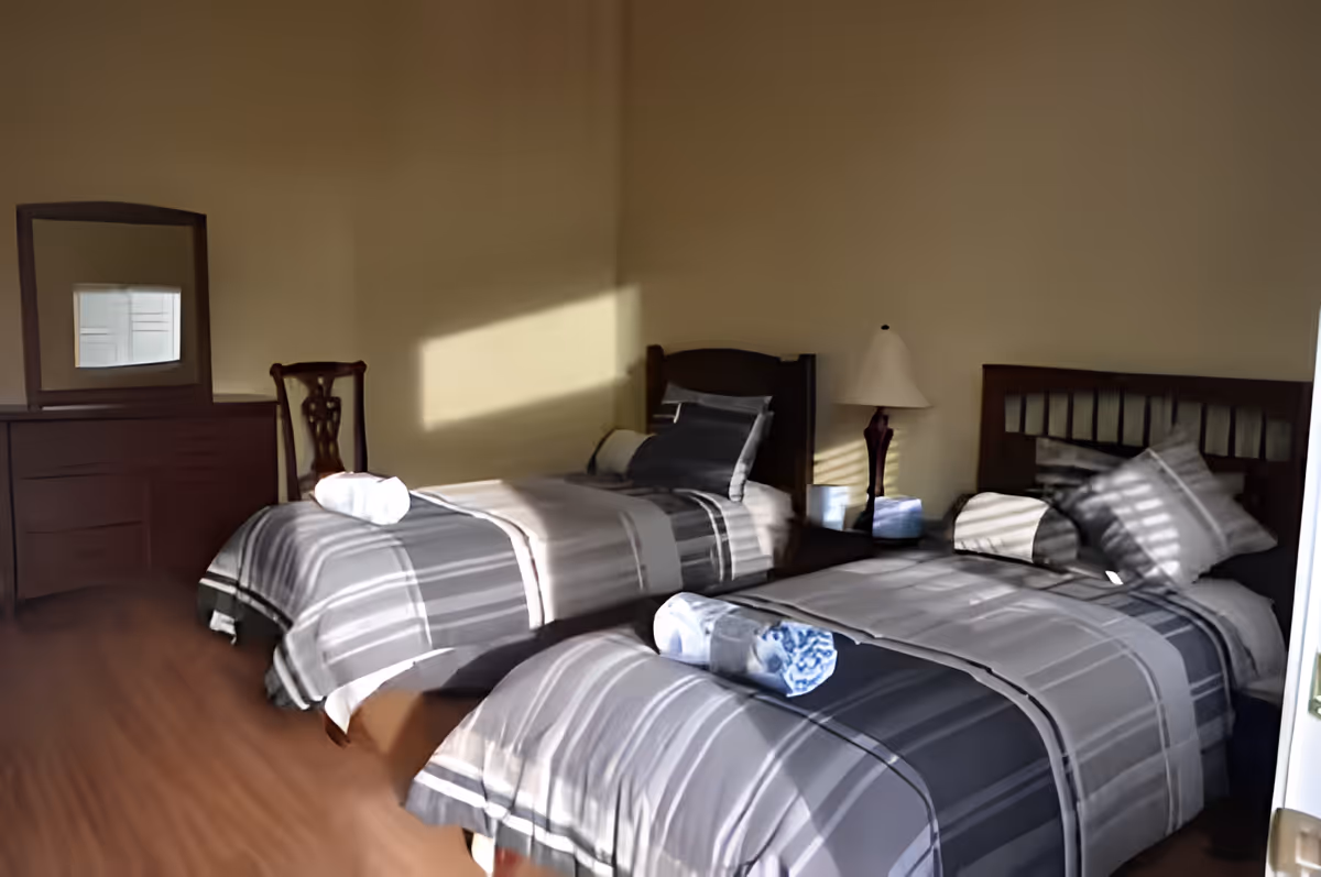 A bedroom with two twin beds, each made with gray and white striped bedding and a rolled towel placed on top. There is a wooden dresser with a mirror and a wooden chair next to it. A bedside table with a lamp is positioned between the two beds. Sunlight streams through a window, casting shadows on the beds and walls.