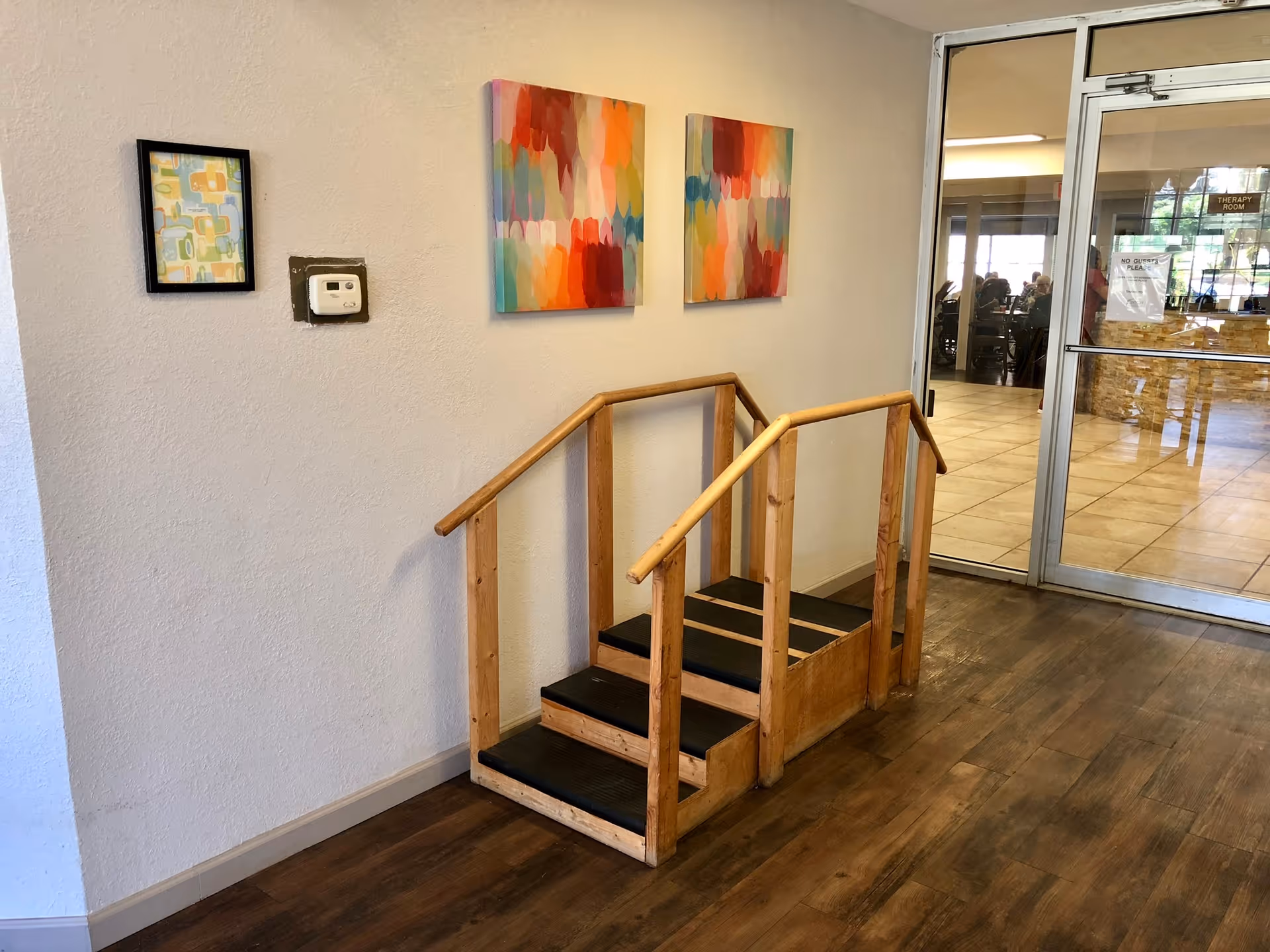 Interior view of a nursing center hallway with wooden handrails and steps for mobility assistance. The wall has three colorful abstract paintings and a thermostat. A glass door leads to a room with people seated inside.