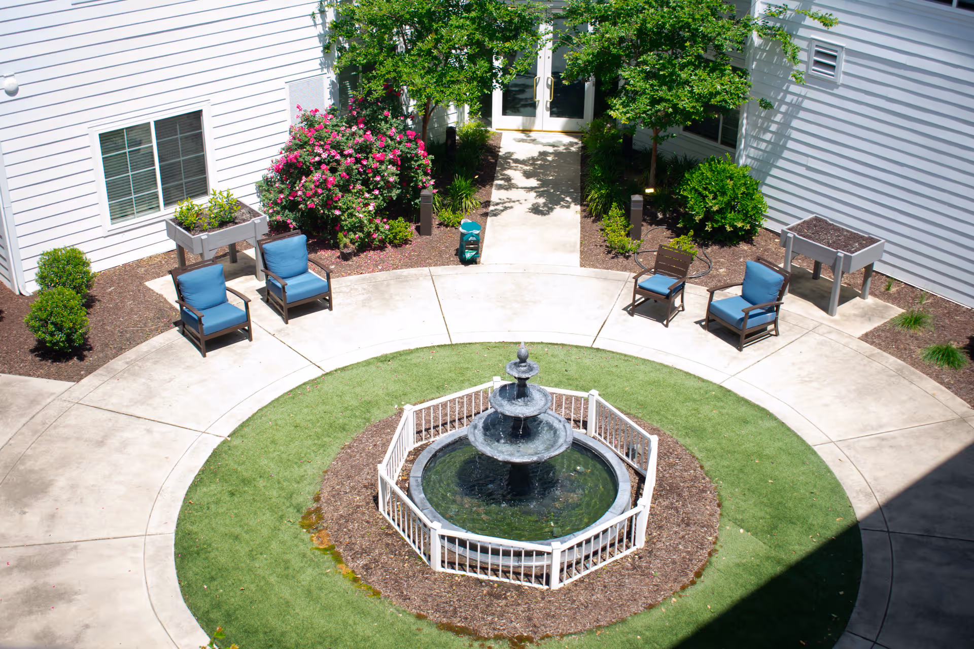 Outdoor courtyard area with a three-tiered water fountain surrounded by a small white fence. The courtyard has a circular concrete walkway with green grass and mulch around the fountain. There are four blue cushioned chairs and two brown chairs placed around the walkway. The area is bordered by white building walls with windows, green bushes, flowering plants, and two raised garden beds.